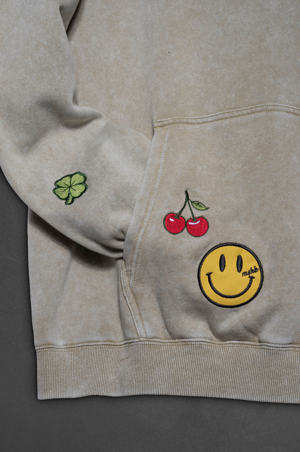 Detalle del bordado cerezas y carita feliz en bolsillo del hoodie khaki